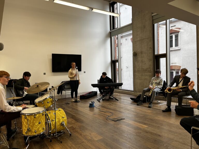 Young musicians from Jazzlife Juniors at rehearsal. In the picture you can see a young man playing drums, a young man playing bass guitar, a young woman singing, a young man playing keyboard, a young man holding a clarinet and a young man holding a saxaphone. In the corner of the picture you can just see the hands of the conductor.