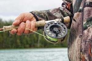 Close up shot of a man holding a fishing rod with a bright green line. IN a blurred background you can see trees and a body of water. 