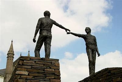 Maurice Harron's Hands Across the Divide piece in Derry/Londonderry. A bronze monument featuring two larger-than-life men standing on separate, curved stone bases. They lean slightly forward, reaching out towards one another across a physical gap. Their fingertips are close but do not touch, symbolising a hopeful journey toward peace, reconciliation, and future connection.